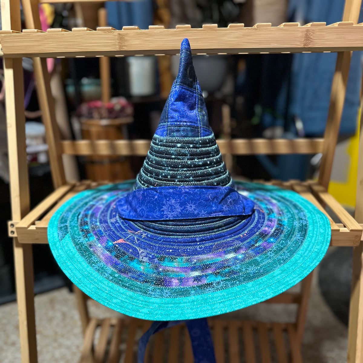 A wizard hat with a wide brim featuring a blue and purple galaxy print, displayed on a wooden stand.