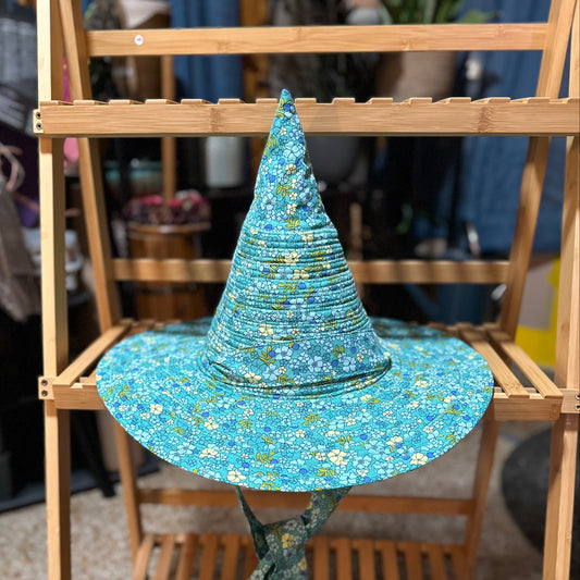 A wide-brimmed witch hat in a blue and green floral print fabric, displayed on a wooden shelf with a matching fabric chin strap.