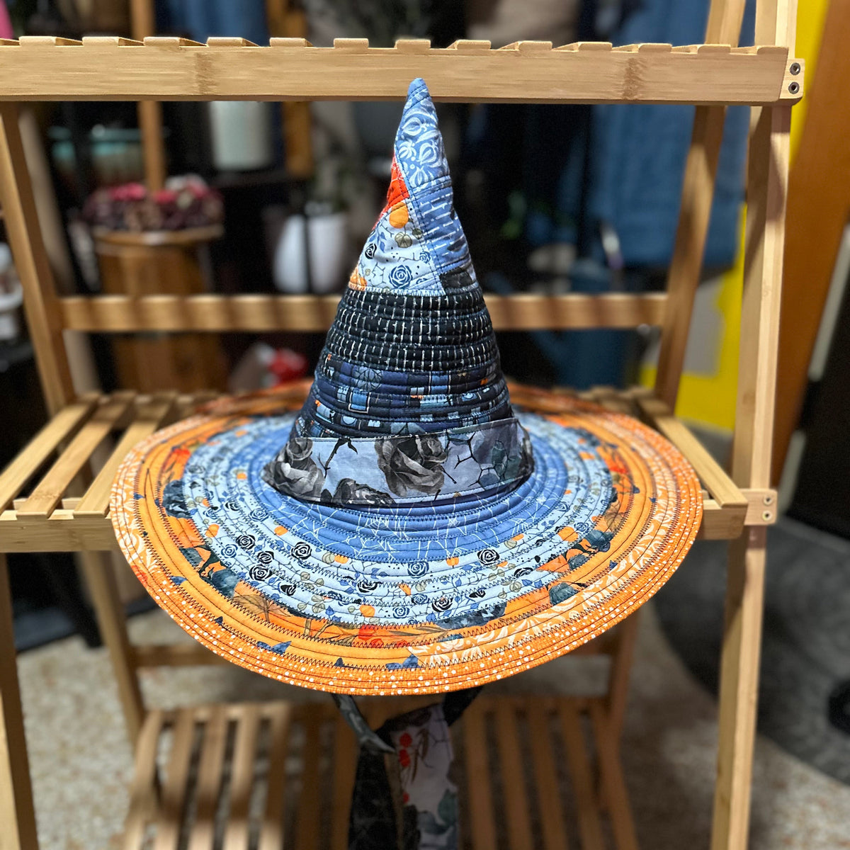 A witch-style pointed hat with a wide brim, displayed on a wooden stand. The hat features a blue and black patterned patterned fabric body with a bright orange brim decorated with small white and yellow motifs, and a gray patterned ribbon hanging from the bottom.