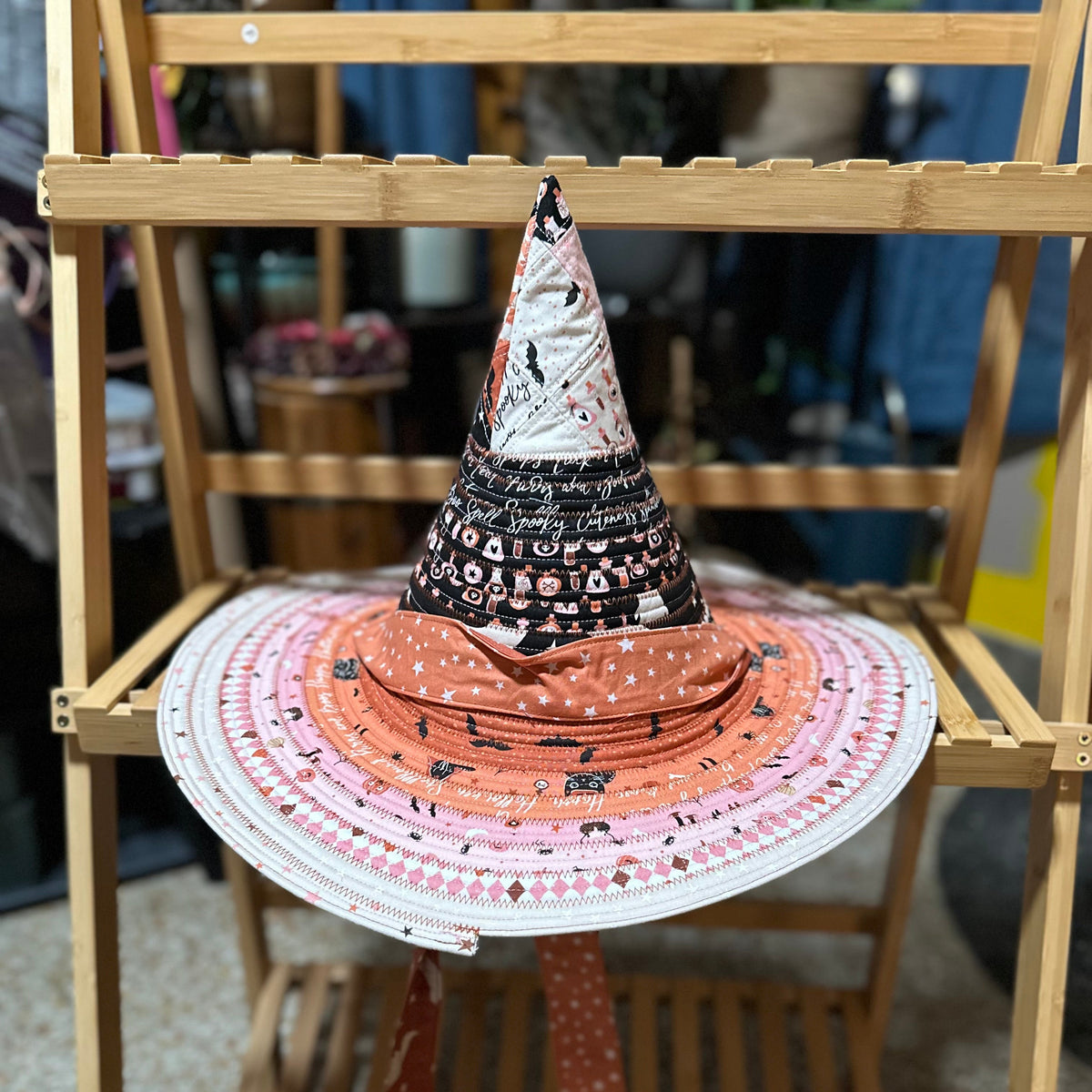 A wide-brim sun hat with a witch hat topper, displayed on a wooden shelf. The hat features a pink and white patterned brim with black spider motifs, an orange crown, and a black pointed witch hat with white text.