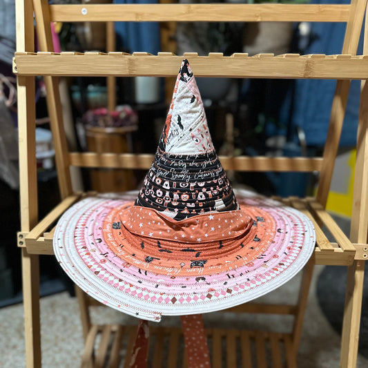 A wide-brim sun hat with a witch hat topper, displayed on a wooden shelf. The hat features a pink and white patterned brim with black spider motifs, an orange crown, and a black pointed witch hat with white text.
