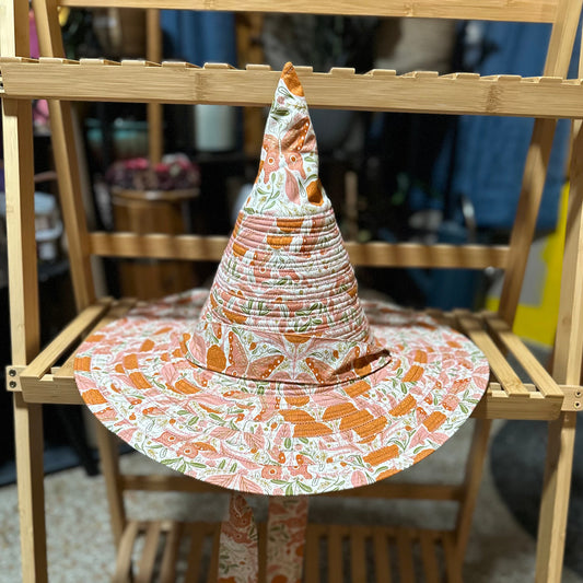 A pointed witch hat with a wide brim, featuring an orange, pink, and green patterned fabric, displayed on a wooden folding stand.