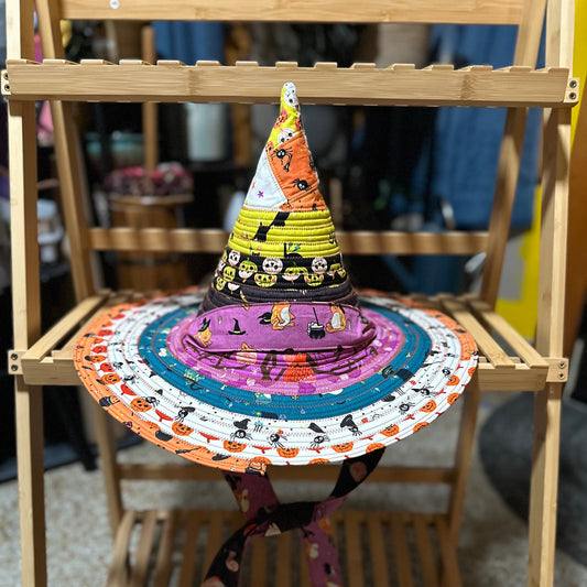 A handmade multicolor fabric witch hat with a wide brim, displayed on a wooden folding chair. The hat features a pointed yellow and black patterned cone top, a purple brim with a black witch hat print, and a black chin strap with orange and black Halloween motifs.